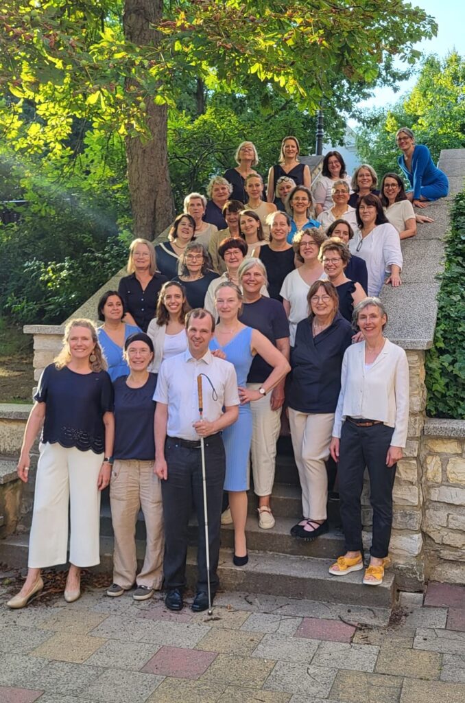 Chorfoto im Freien auf einer Treppe, im Hintergrund Büsche, Bäume und blaer Himmel, etwa 30 Frauen und ein Mann, alle in den Farben Blau, Beige und Weiß gekleidet